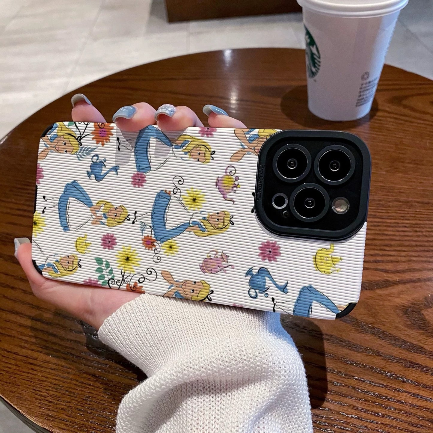 iPhone case with Alice in the Wonderland design held by a hand on a wooden table with a Starbucks cup in the background