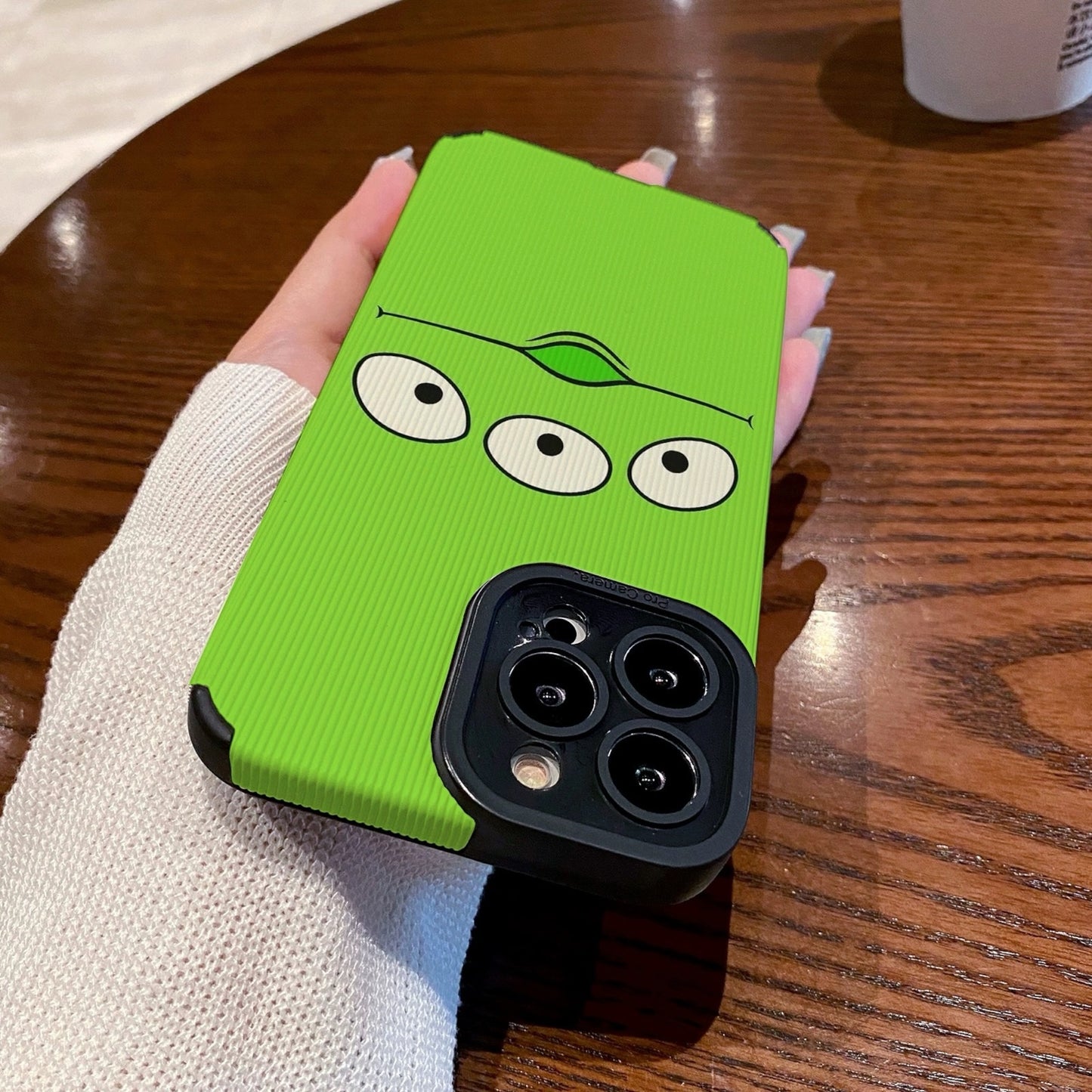Person holding a green toy story alien phone case with cartoon face design on a wooden table with a Starbucks cup.
