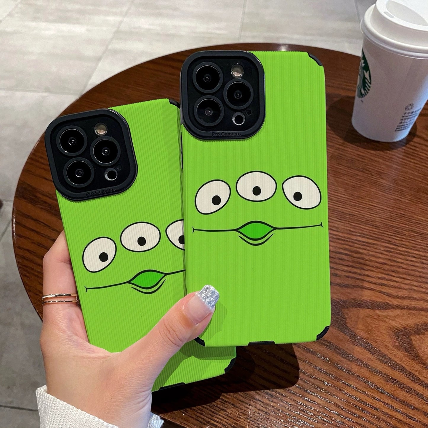 Person holding a green toy story alien phone case with cartoon face design on a wooden table with a Starbucks cup.