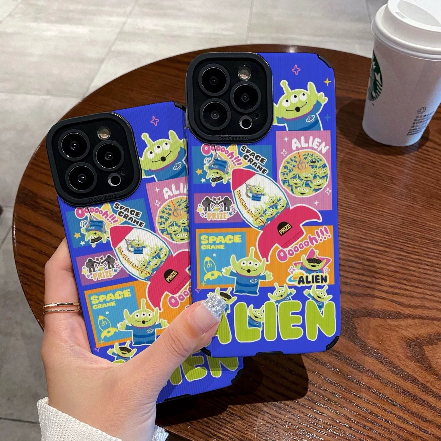 Colorful phone case with Toy Story alien design held by a person on a wooden table with a Starbucks cup in the background.