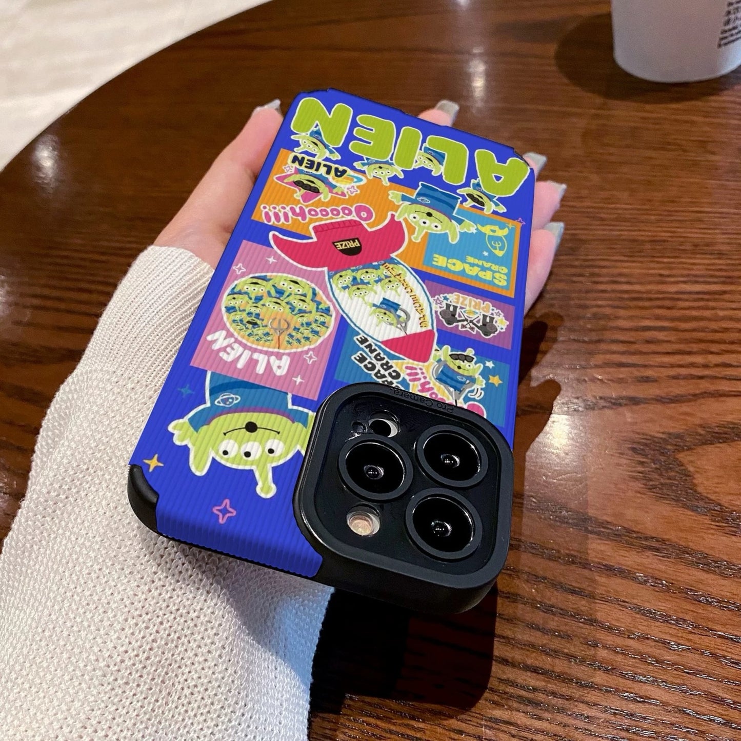 Colorful phone case with Toy Story alien design held by a person on a wooden table with a Starbucks cup in the background.