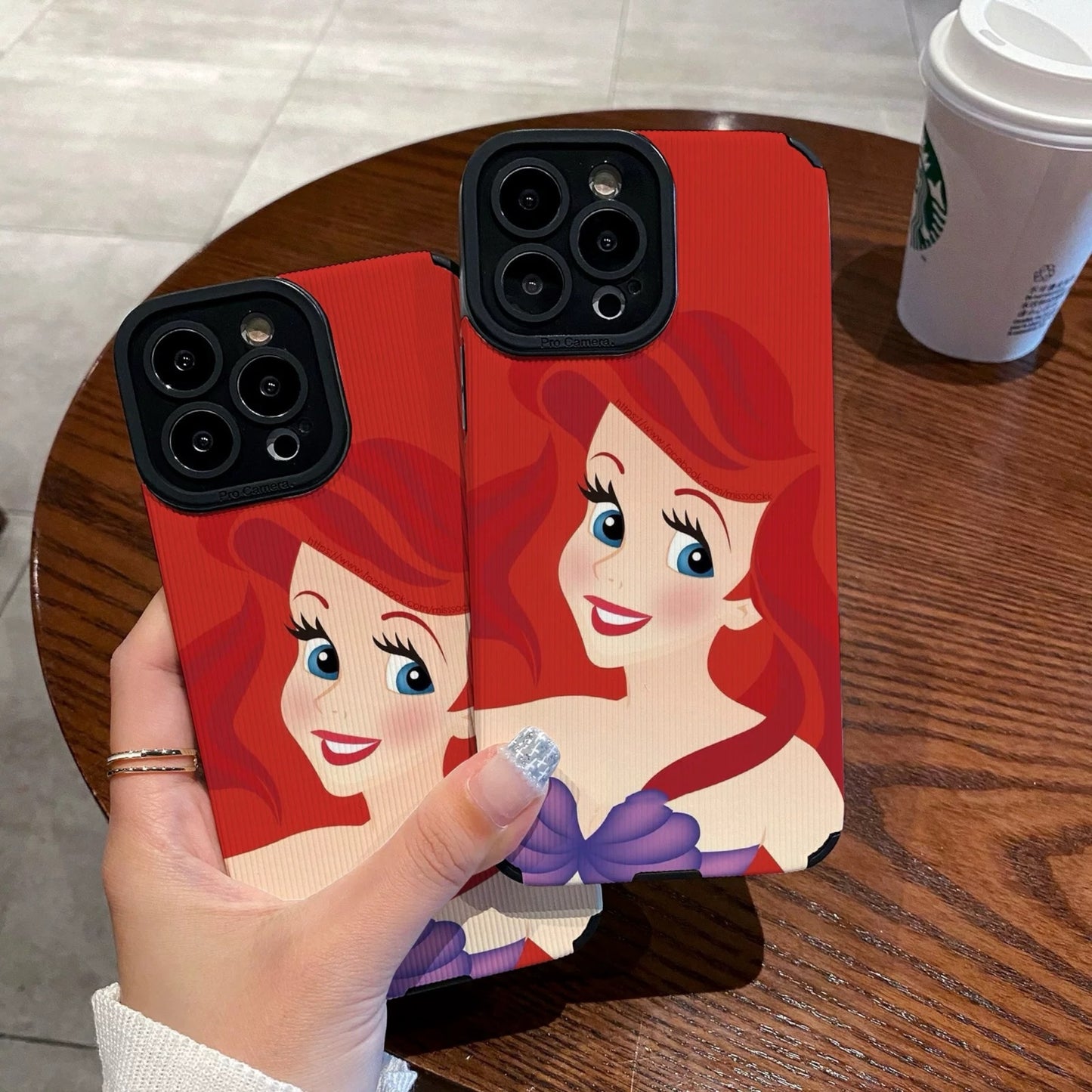 My Little Mermaid Ariel iPhone case with a character design held by a hand on a wooden table with a Starbucks cup in the background.