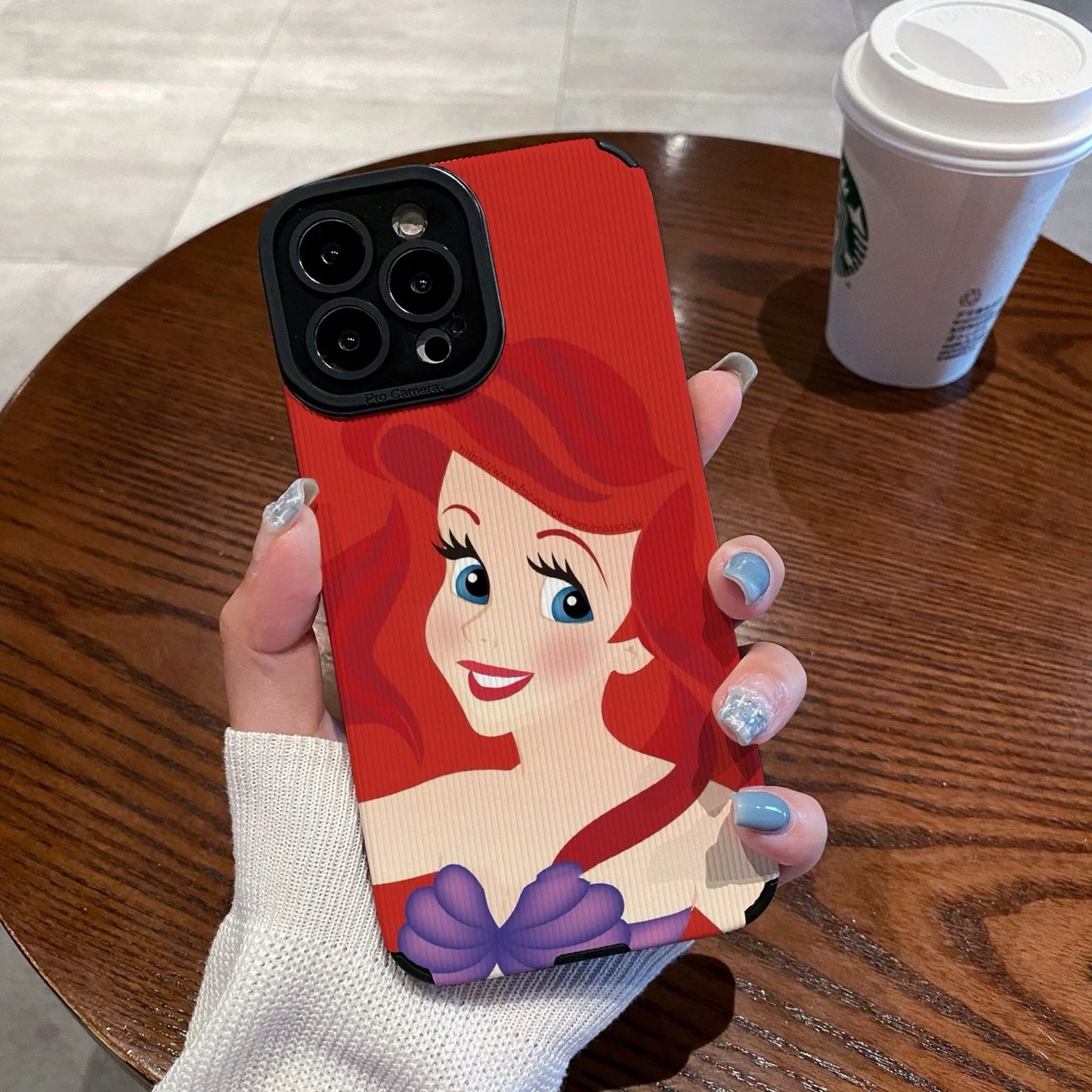 My Little Mermaid Ariel iPhone case with a character design held by a hand on a wooden table with a Starbucks cup in the background.