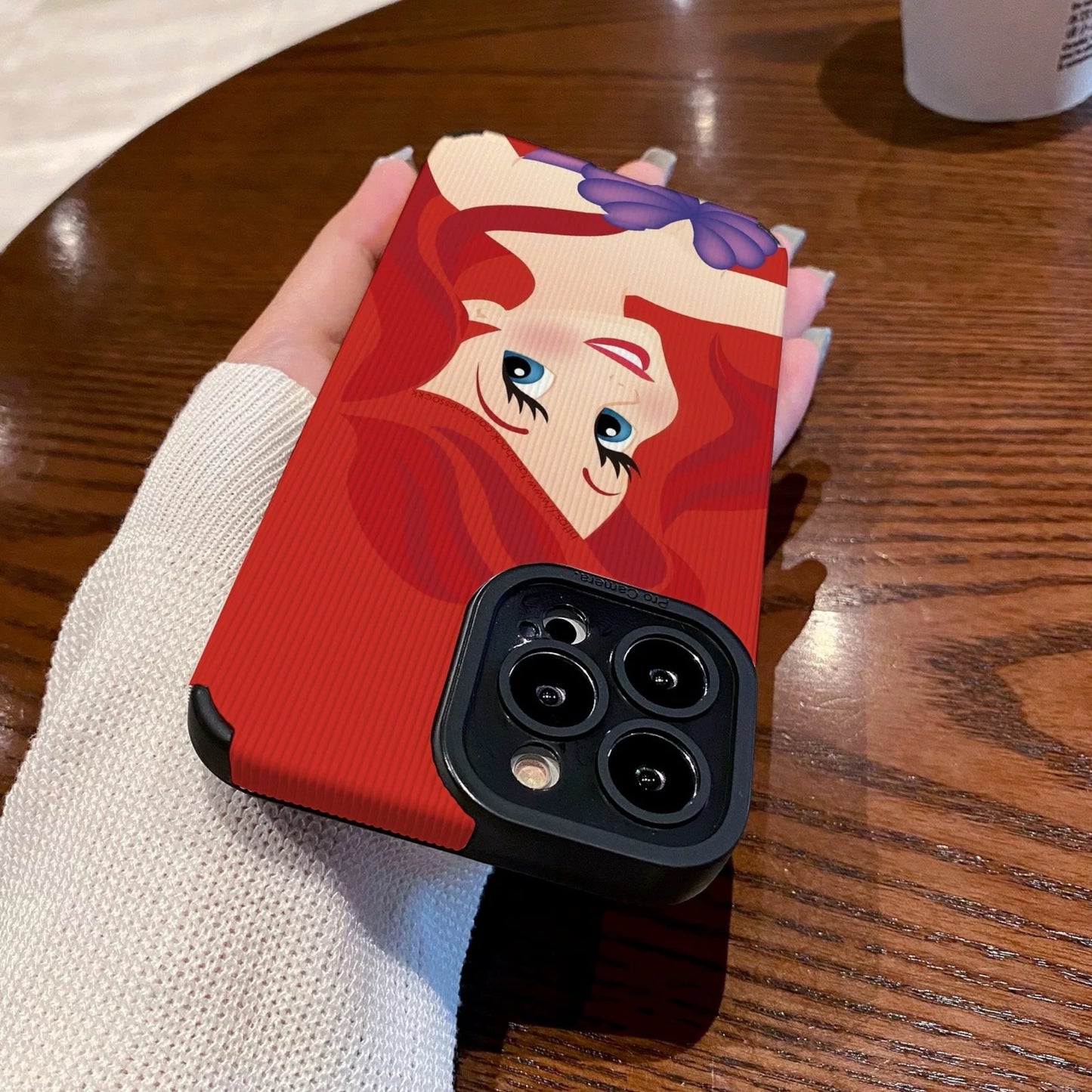 My Little Mermaid Ariel iPhone case with a character design held by a hand on a wooden table with a Starbucks cup in the background.