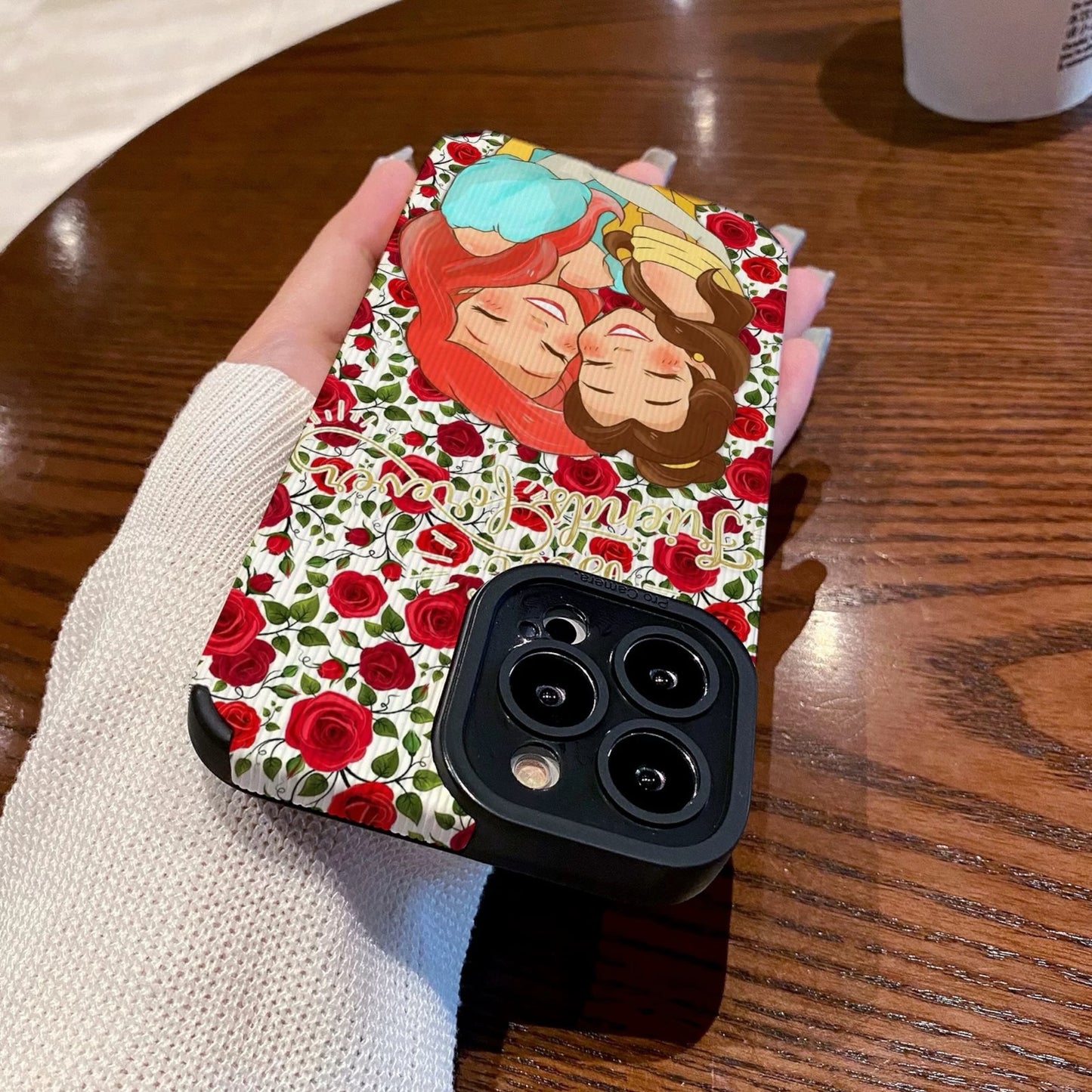 iPhone case with cartoon characters Princess Ariel and Belle on a wooden table with a Starbucks cup.