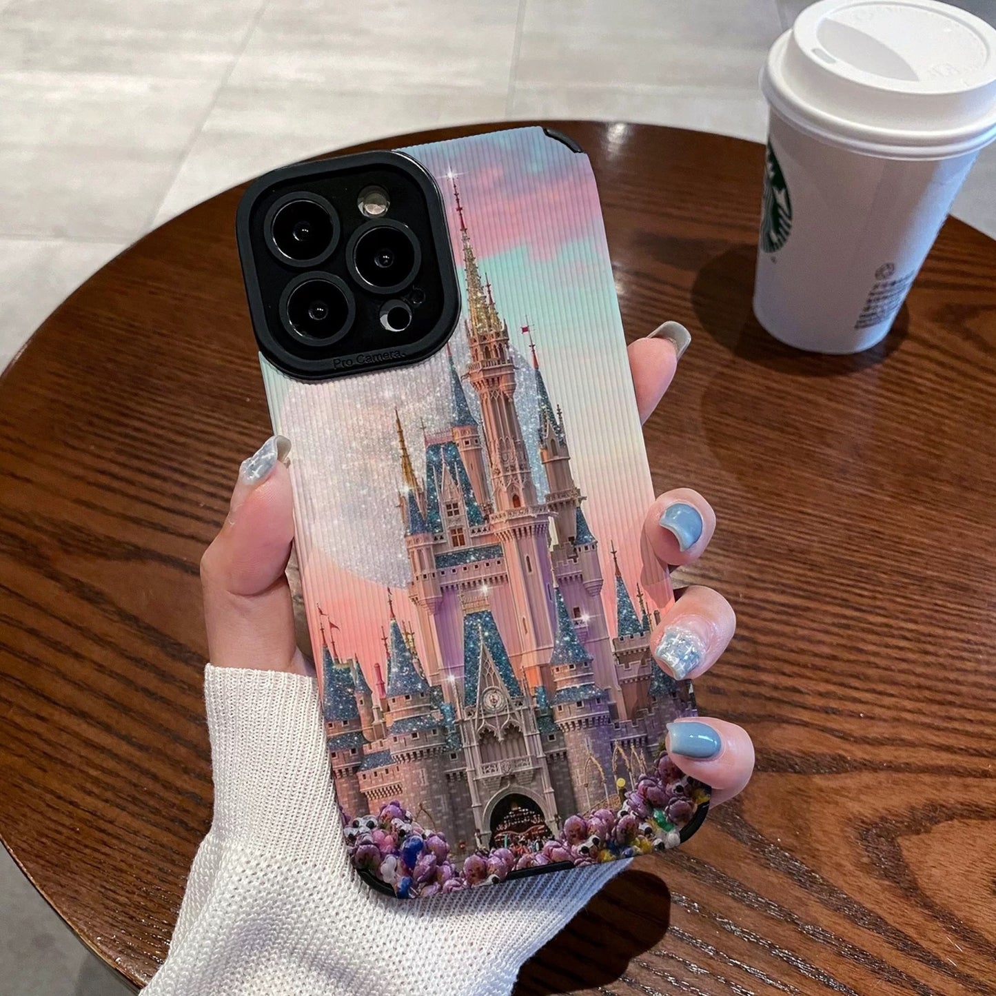 Person holding a iphone with a colorful iphone case featuring a disney theme park castle design, Starbucks cup in the background.