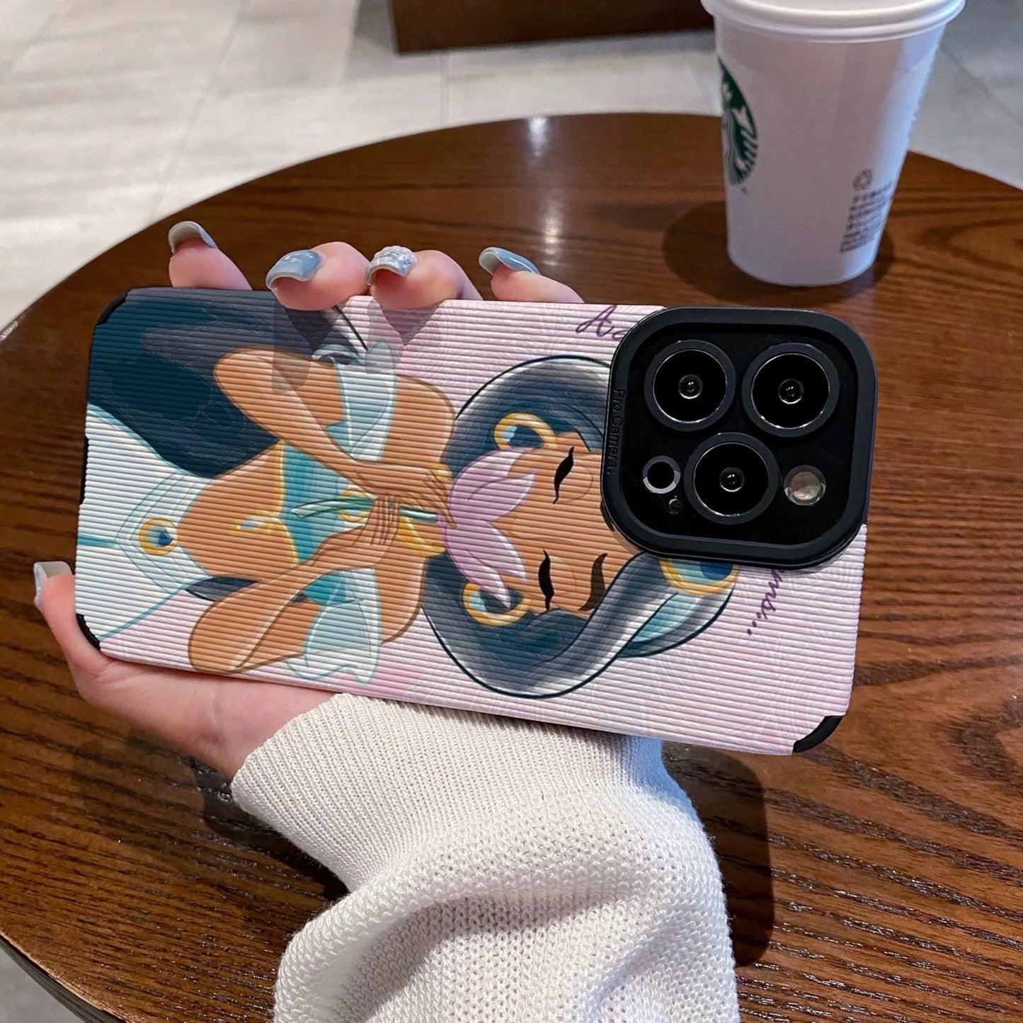 iPhone case with an illustration of a Jasmine holding a flower, held by a hand on a wooden table with a Starbucks cup.