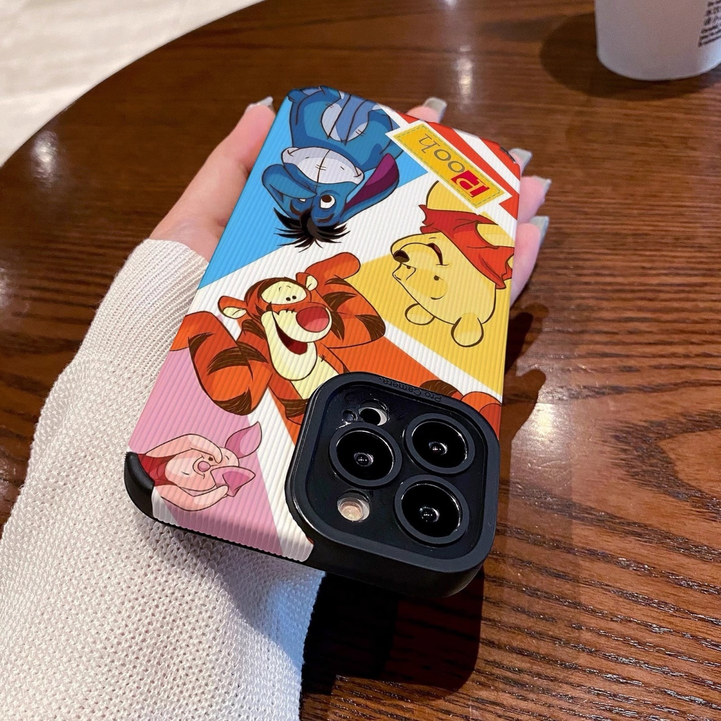 Phone case with cartoon characters winnie the pooh and friends held by a hand on a wooden table with a coffee cup.