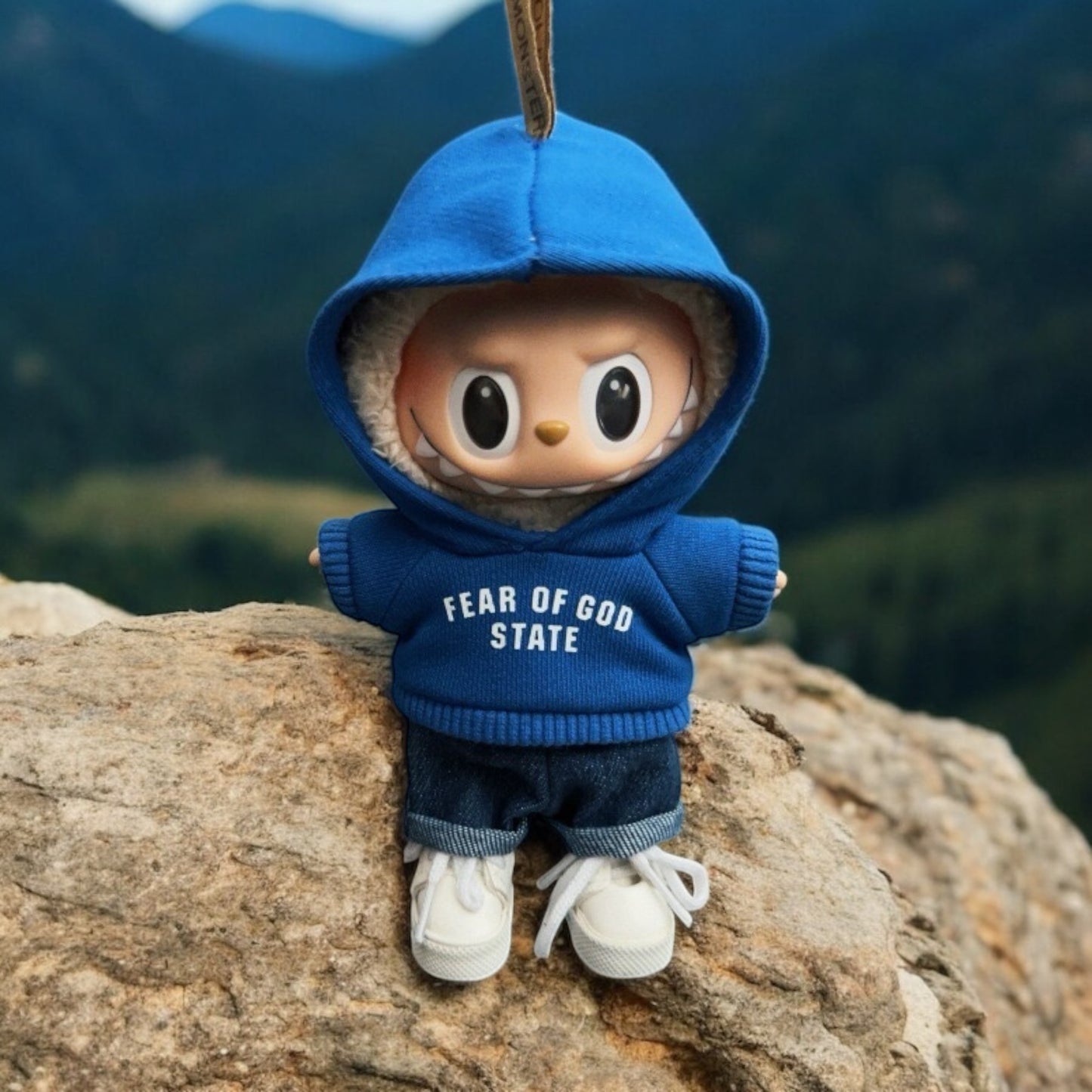 Toy figure Labubu wearing a blue hoodie with 'FEAR OF GOD STATE' text, standing on a rock with a mountainous background.