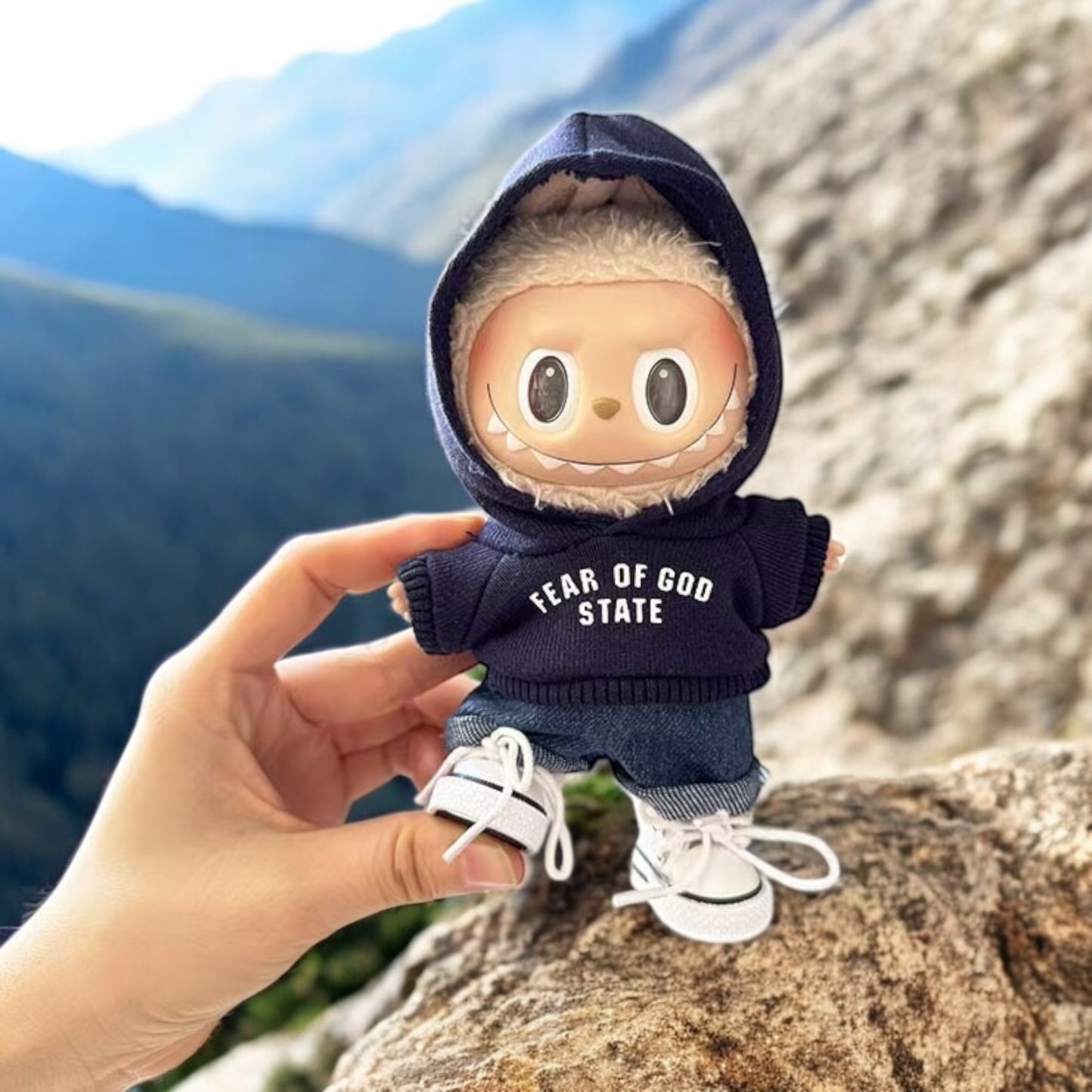Small plush toy Labubu wearing a 'Fear of God State' hoodie held by a hand with a mountainous background
