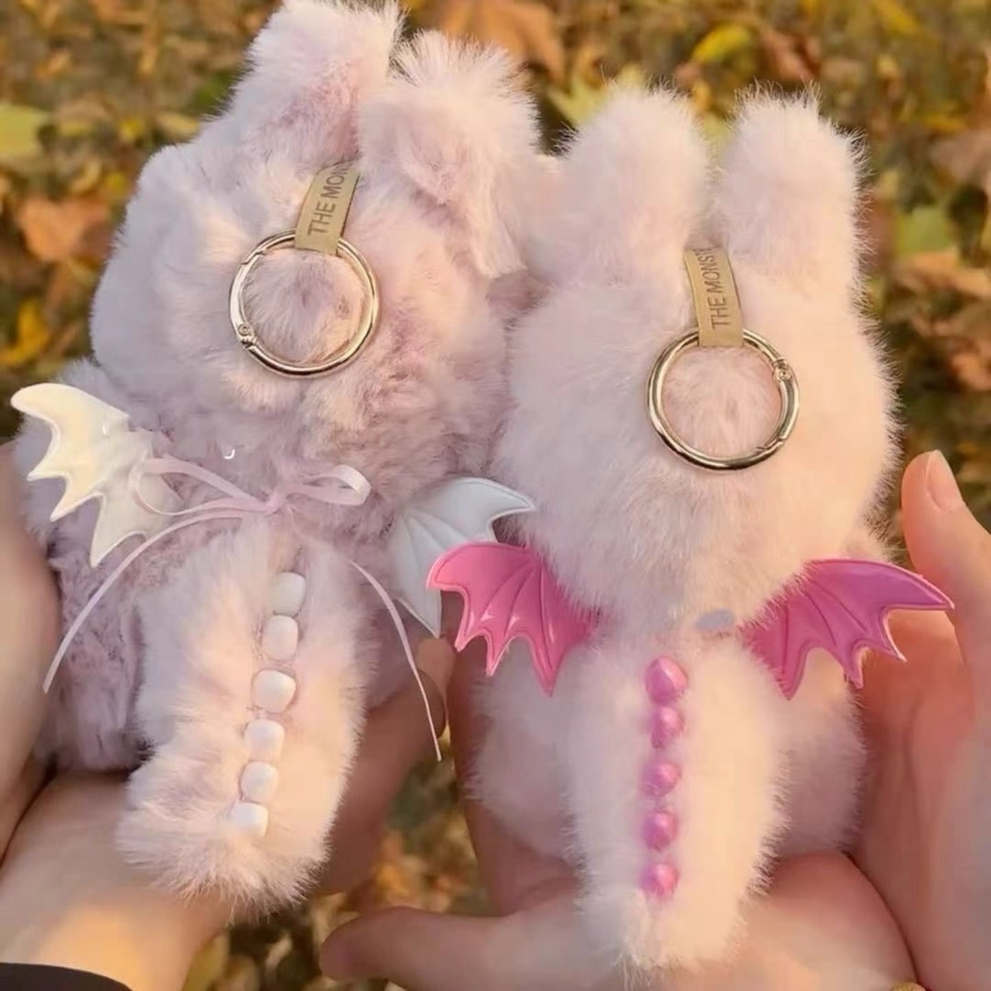 Two Labubu Keychain with fluffy dragon-shaped pink zimomo dinosaurs outfits held by hands with a blurred natural background.