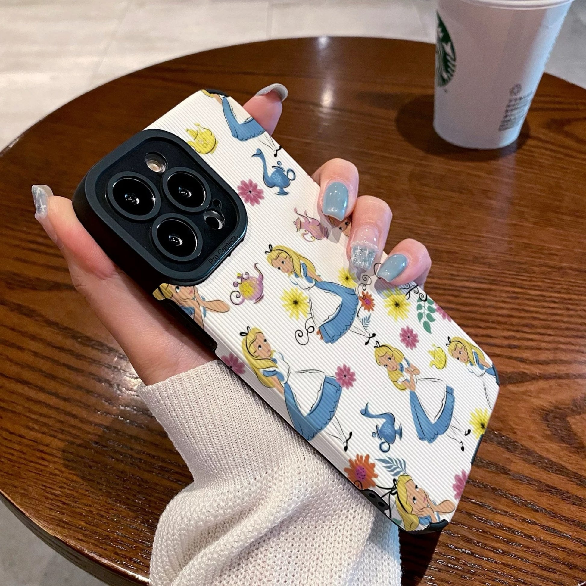 iPhone case with Alice in the Wonderland design held by a hand on a wooden table with a Starbucks cup in the background