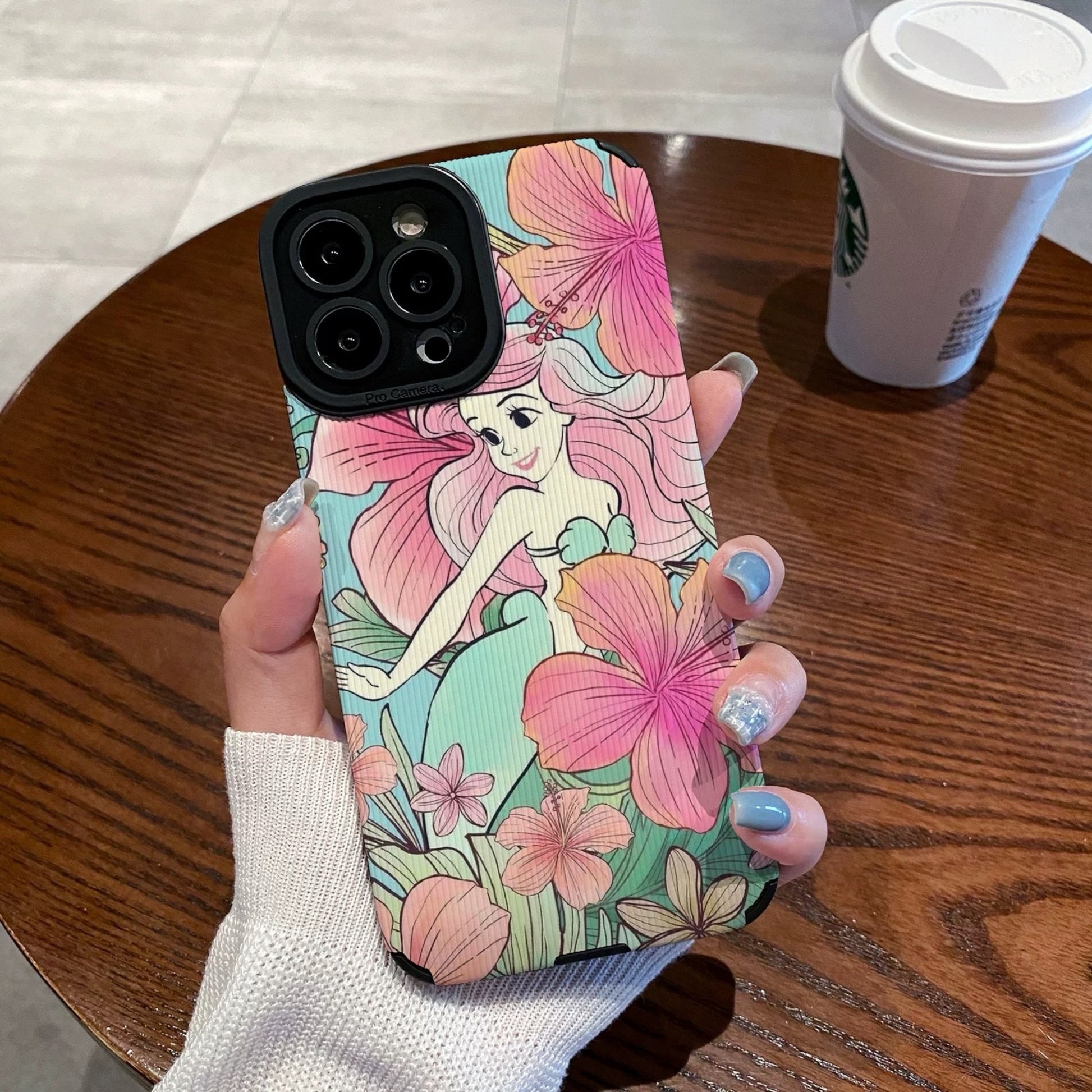 iPhone case with mermaid design held by a hand on a wooden table with a Starbucks cup.