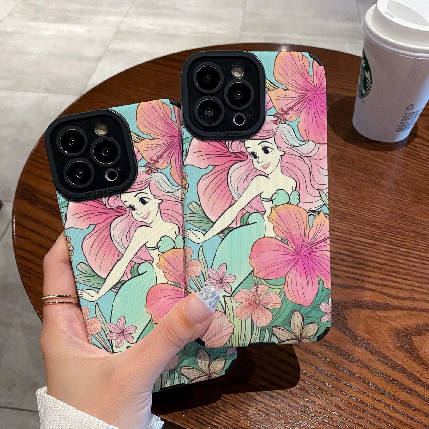 iPhone case with mermaid design held by a hand on a wooden table with a Starbucks cup in the background.