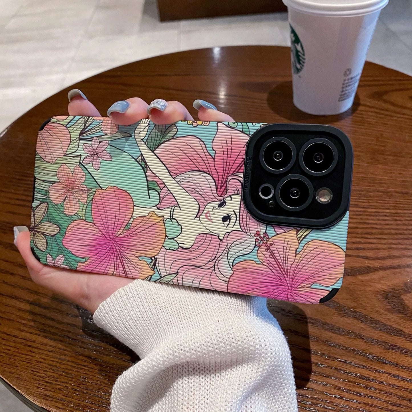 iPhone case with Ariel in  floral design held by a hand on a wooden table with a Starbucks cup in the background.