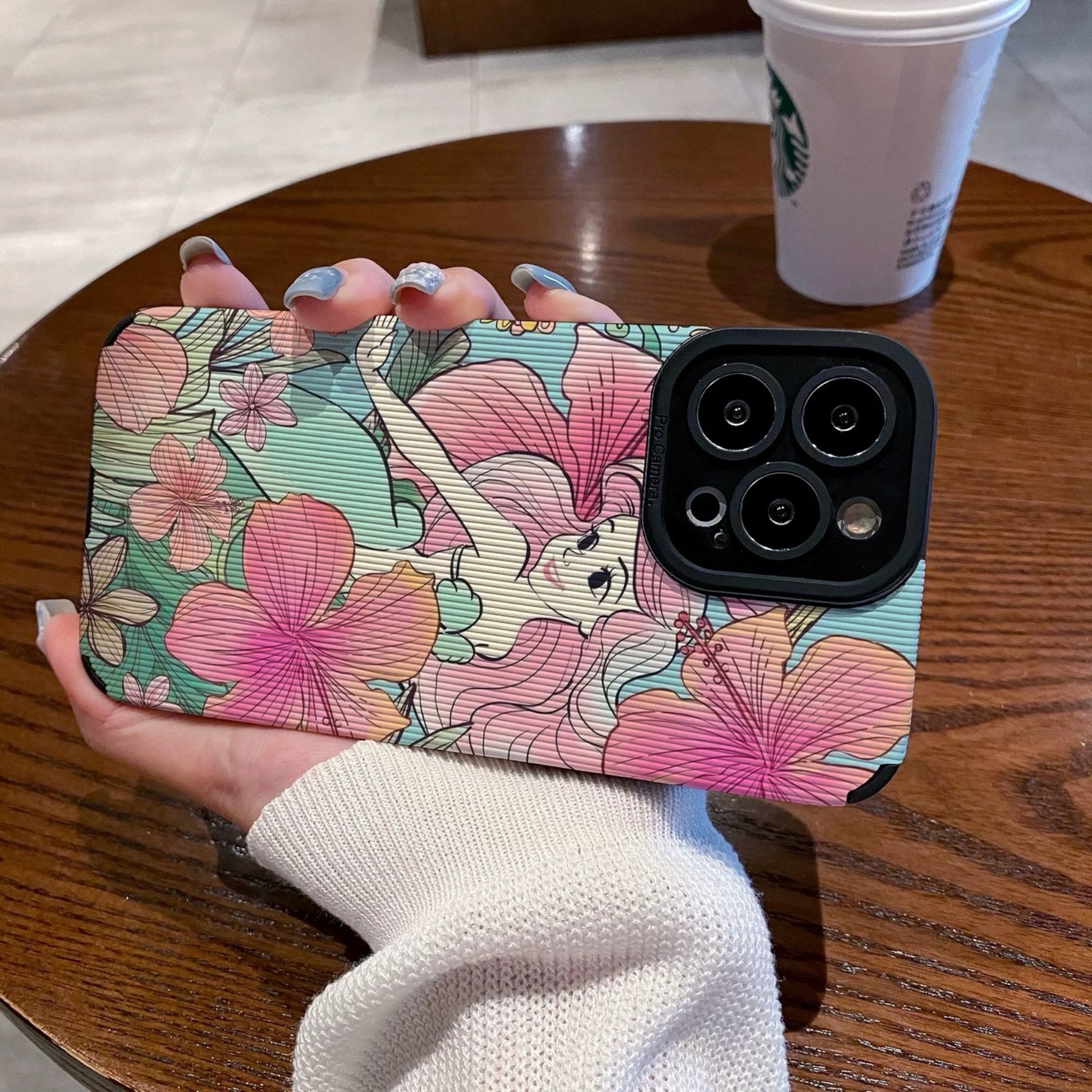 iPhone case with Ariel in  floral design held by a hand on a wooden table with a Starbucks cup in the background.