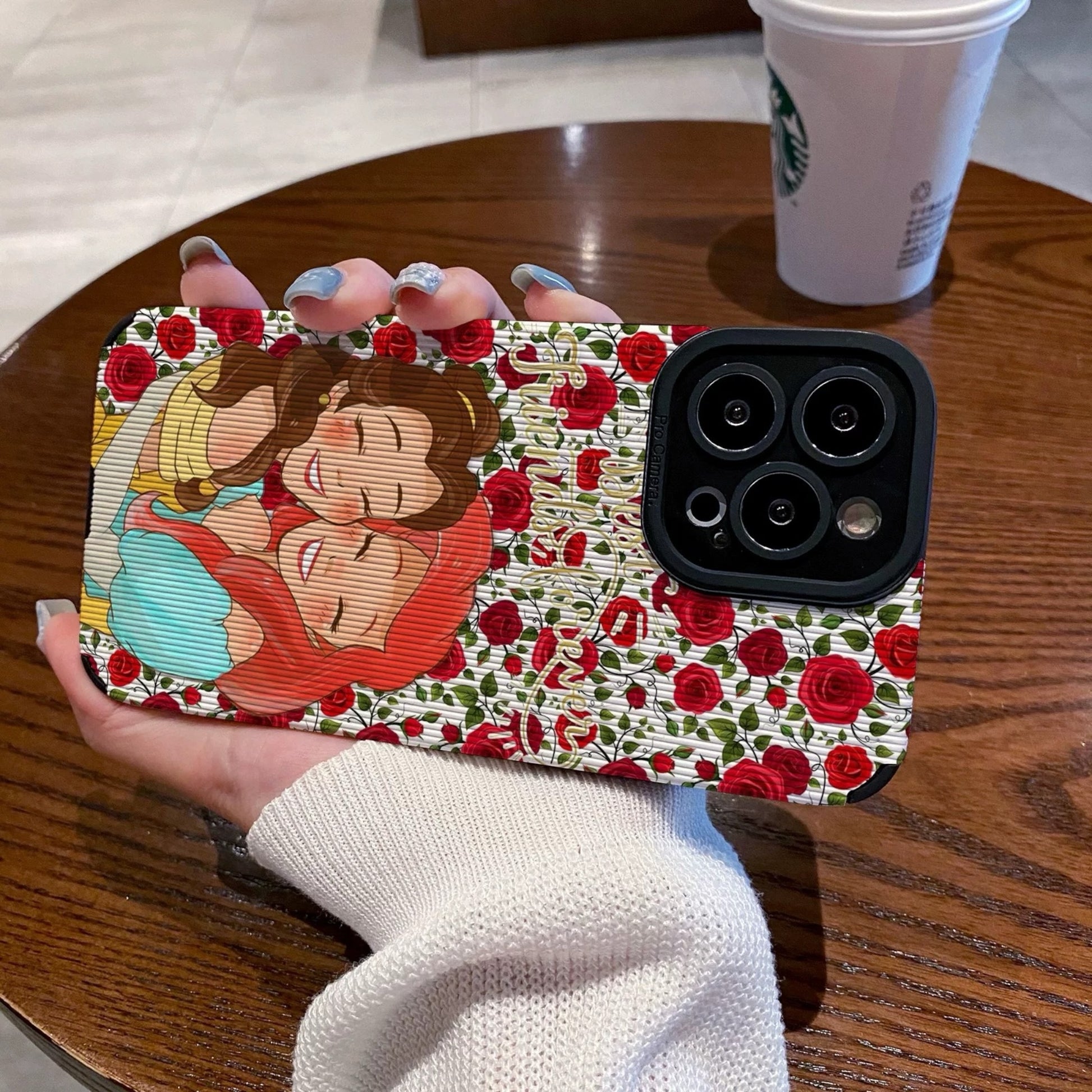 iPhone case with cartoon characters Princess Ariel and Belle on a wooden table with a Starbucks cup.