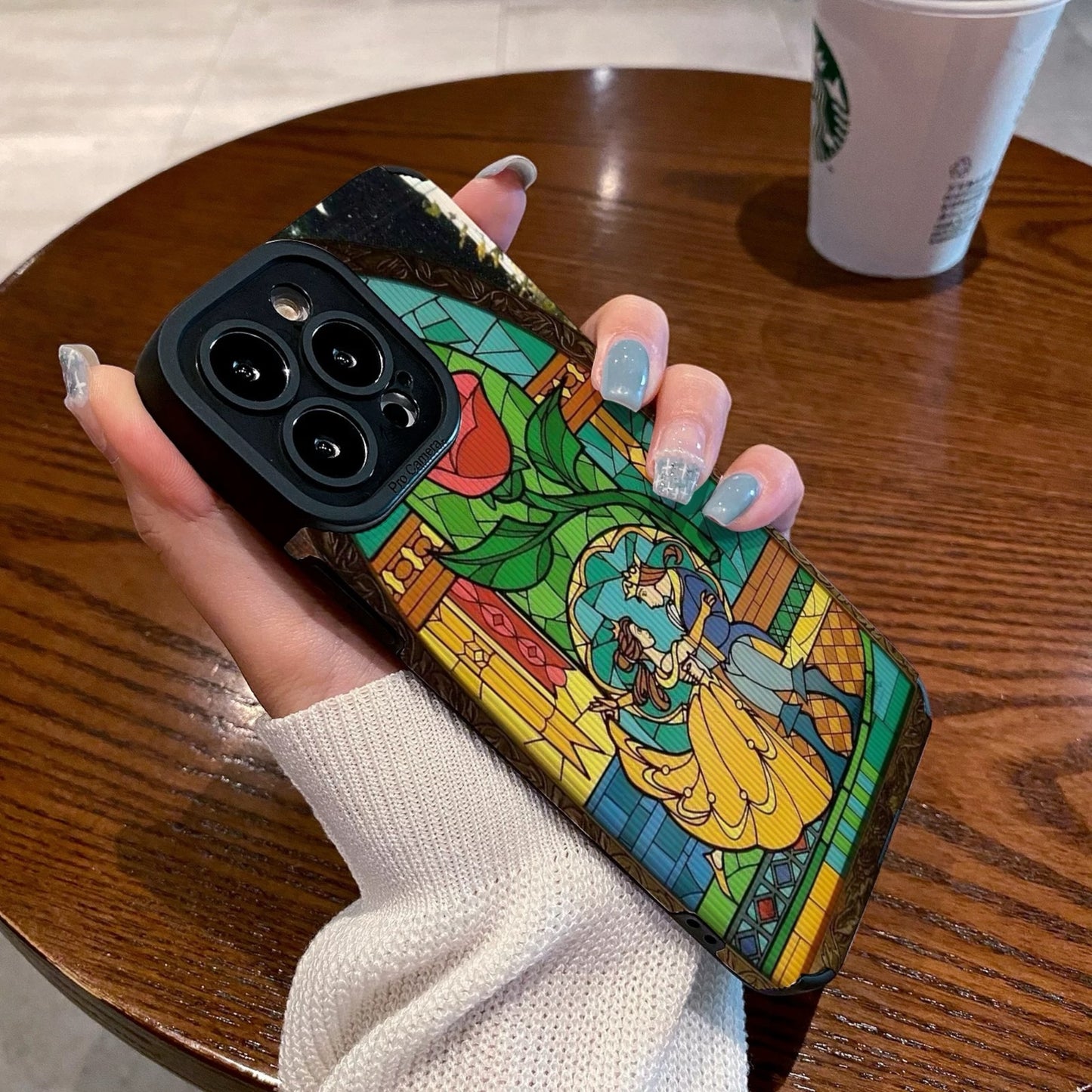 iPhone case with stained glass design with belle and the prince held by a hand on a wooden table with a Starbucks cup.