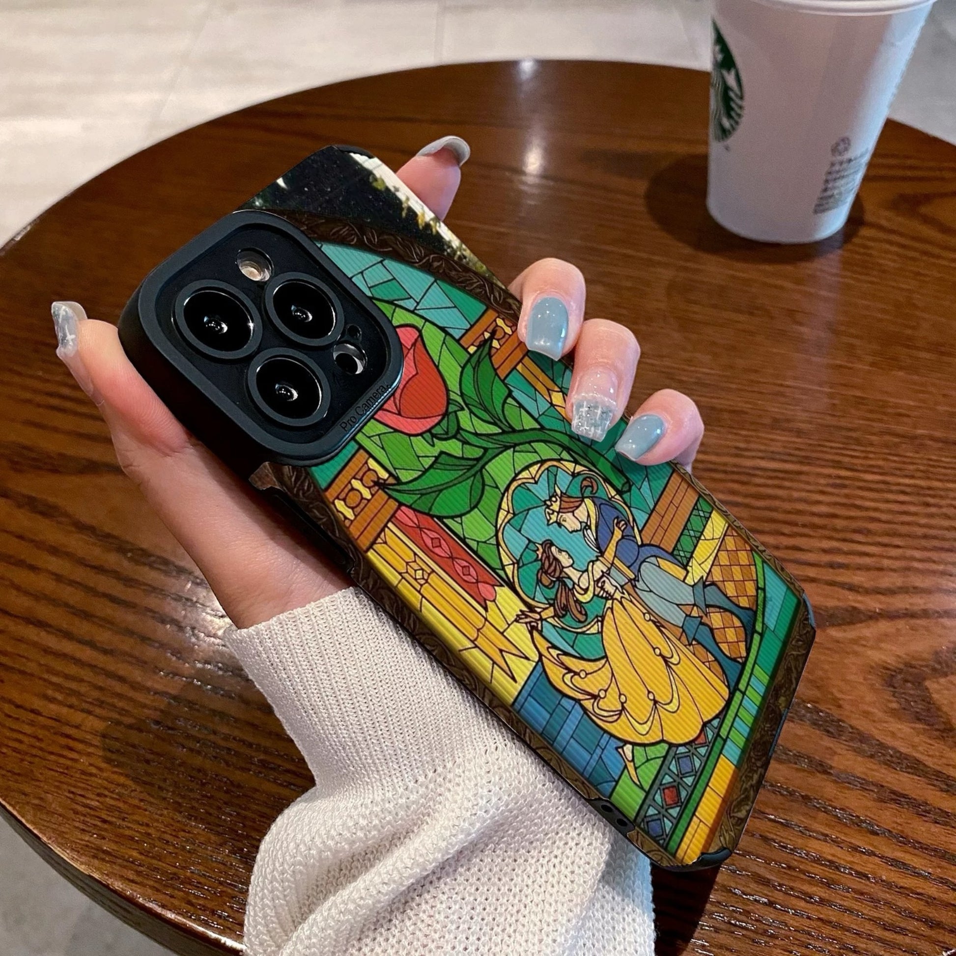 iPhone case with stained glass design with belle and the prince held by a hand on a wooden table with a Starbucks cup.