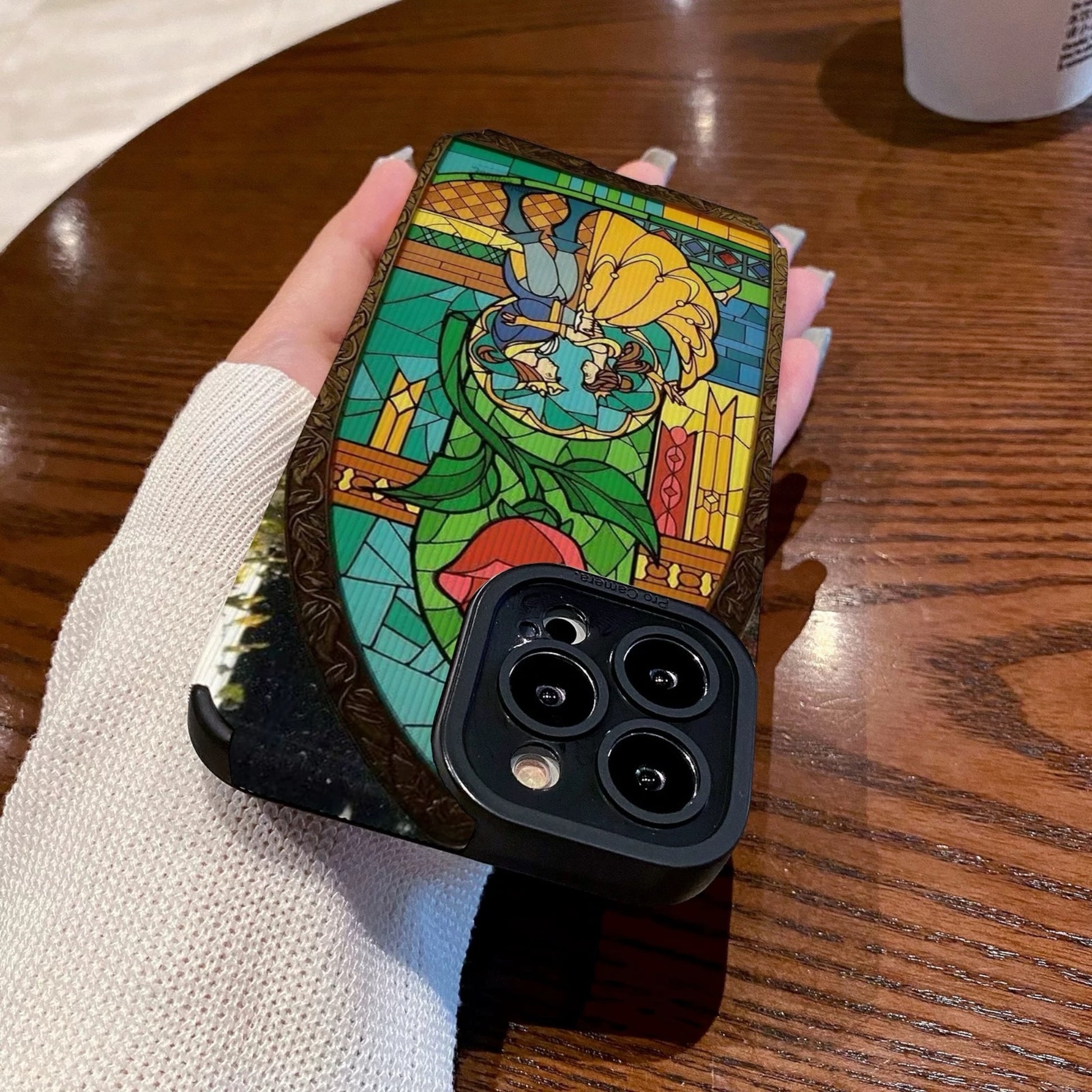 iPhone case with stained glass design with belle and the prince held by a hand on a wooden table with a Starbucks cup.