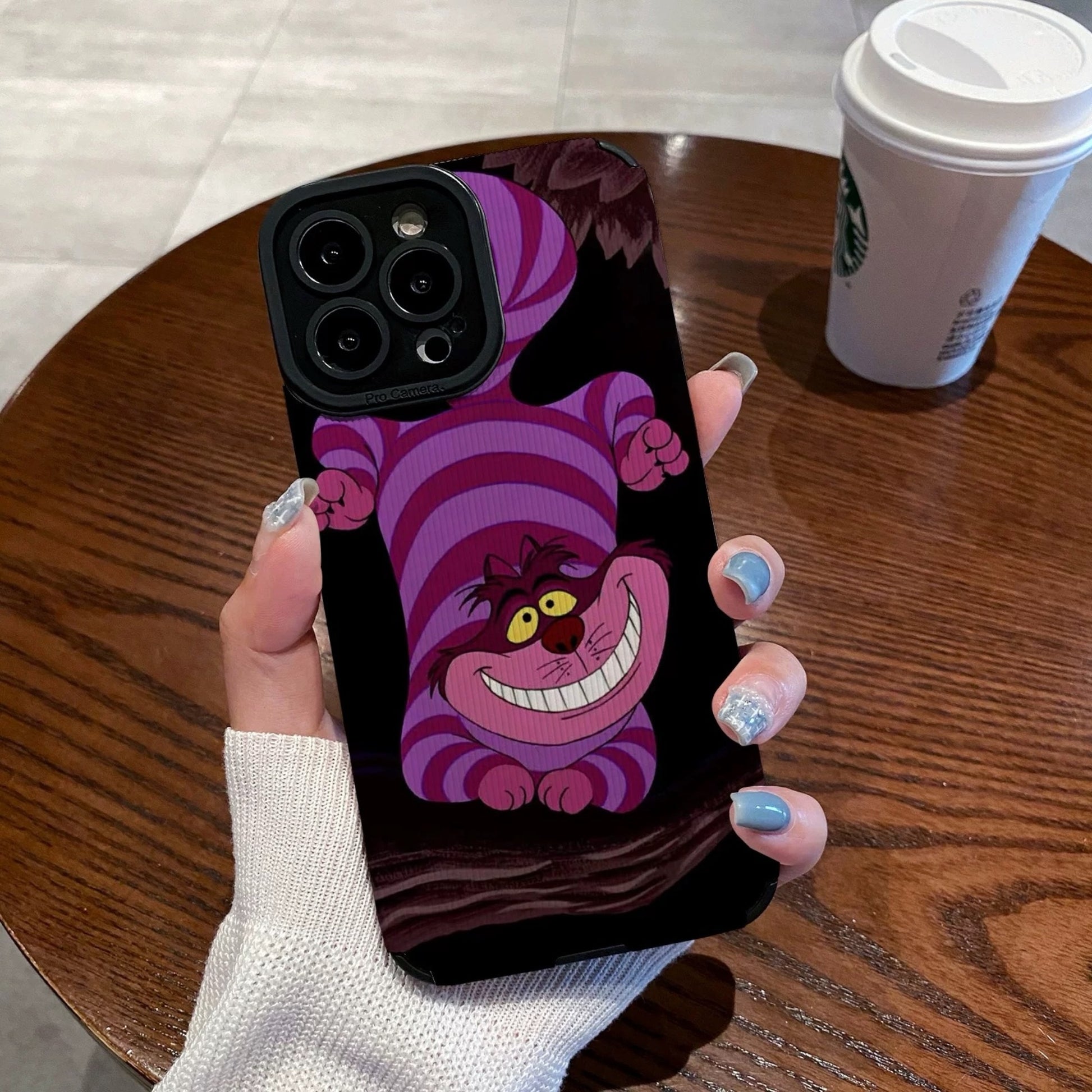 Hand holding a iPhone case with a Cheshire Cat of Alice in Wonderland on a wooden table with a Starbucks cup.