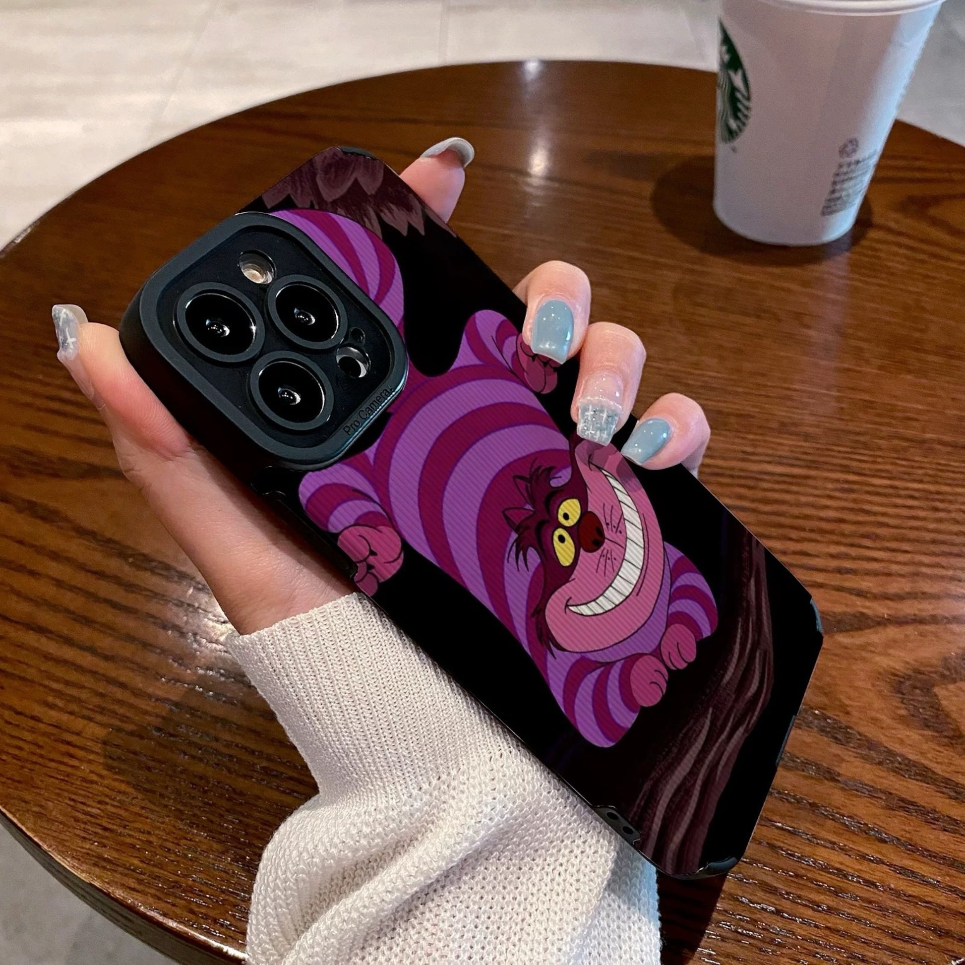 Hand holding a iPhone case with a Cheshire Cat of Alice in Wonderland on a wooden table with a Starbucks cup.
