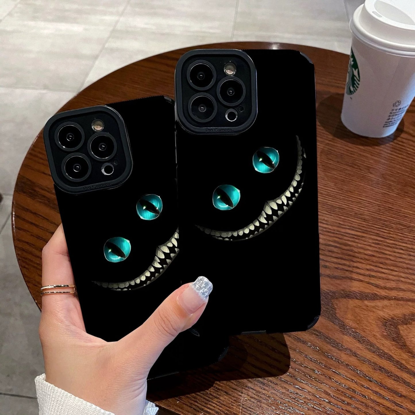 Person holding a black iPhone case with Cheshire Cat of Alice in Wonderland design on a wooden table with a Starbucks cup in the background.