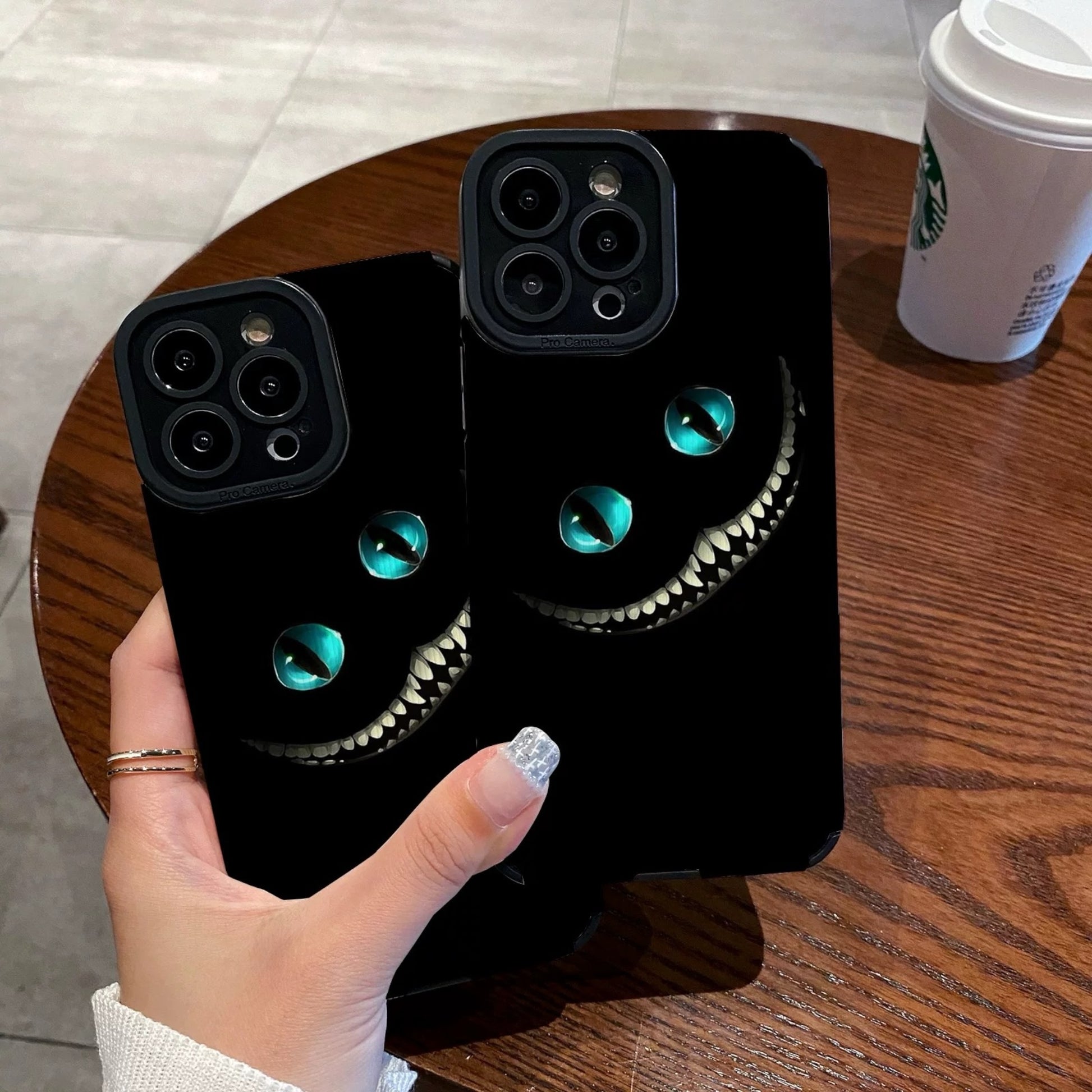 Person holding a black iPhone case with Cheshire Cat of Alice in Wonderland design on a wooden table with a Starbucks cup in the background.