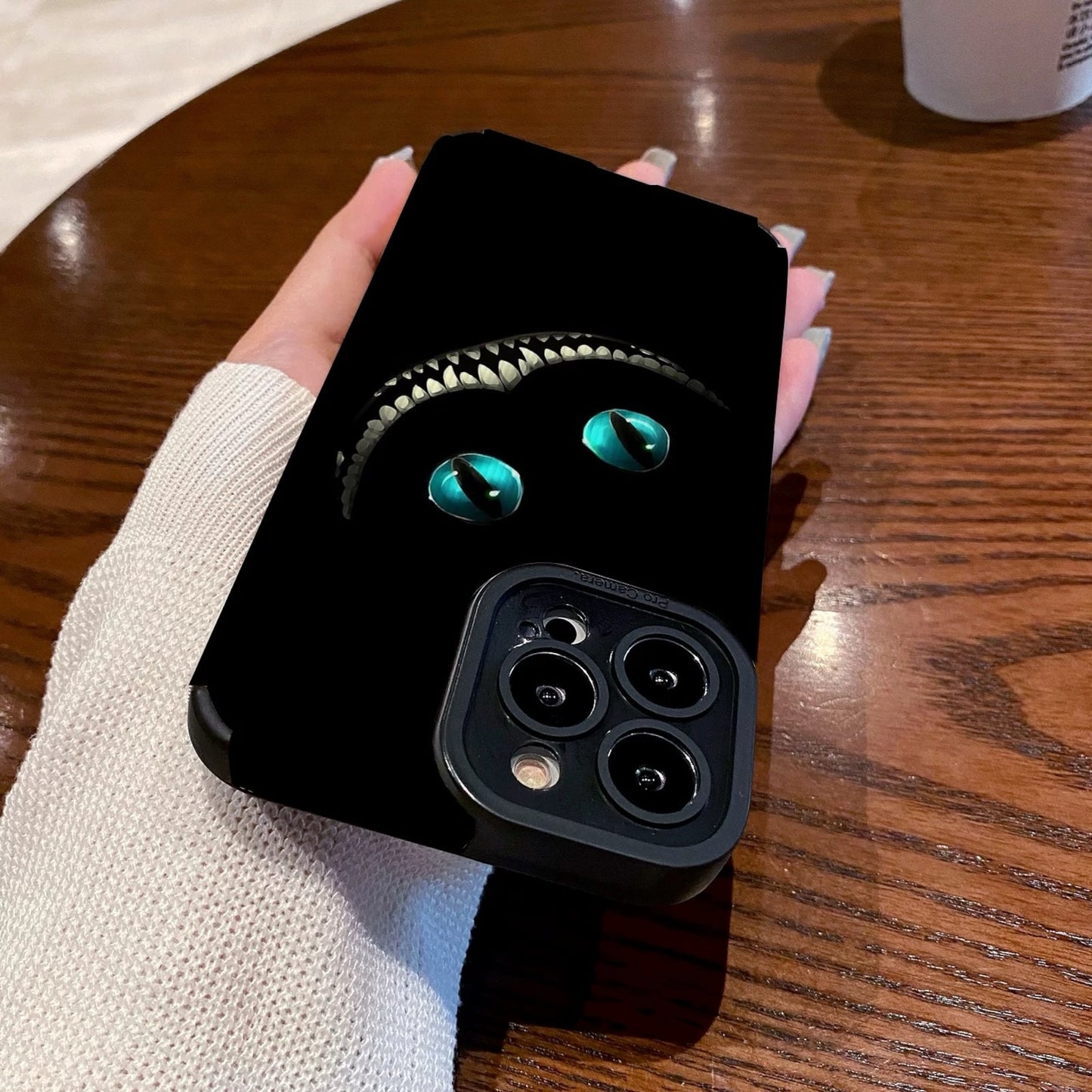 Person holding a black iPhone case with Cheshire Cat of Alice in Wonderland design on a wooden table with a Starbucks cup in the background.