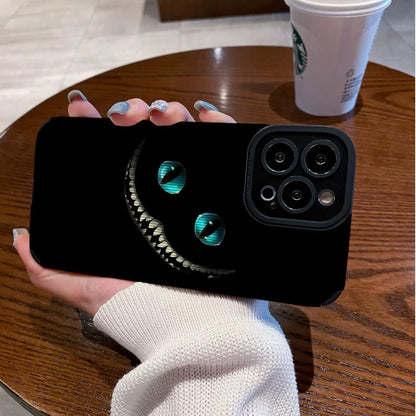 Person holding a black iPhone case with Cheshire Cat of Alice in Wonderland design on a wooden table with a Starbucks cup in the background.