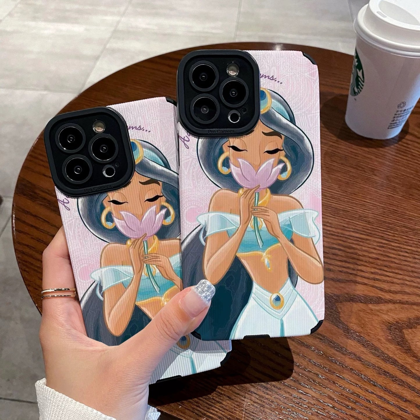iPhone case with an illustration of a Jasmine holding a flower, held by a hand on a wooden table with a Starbucks cup.