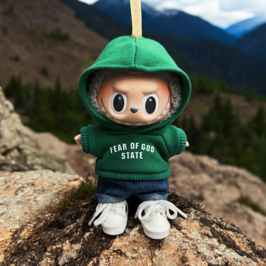 Toy figure Labubu wearing a green hoodie with 'Fear of God State' text, standing on a rock with a mountainous background.