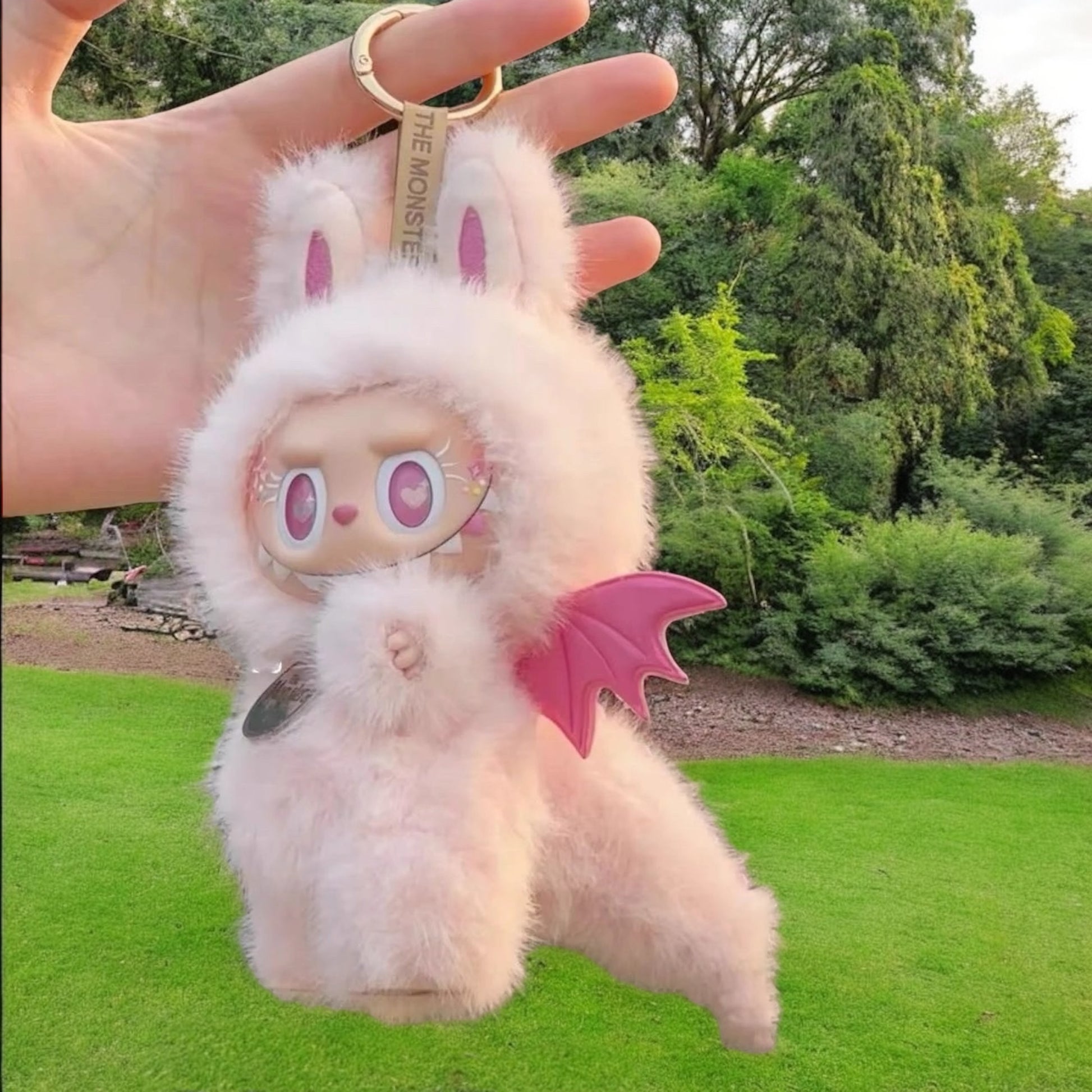 Furry plush keychain with pink zimomo dinosaurs outfit held by a hand outdoors.
