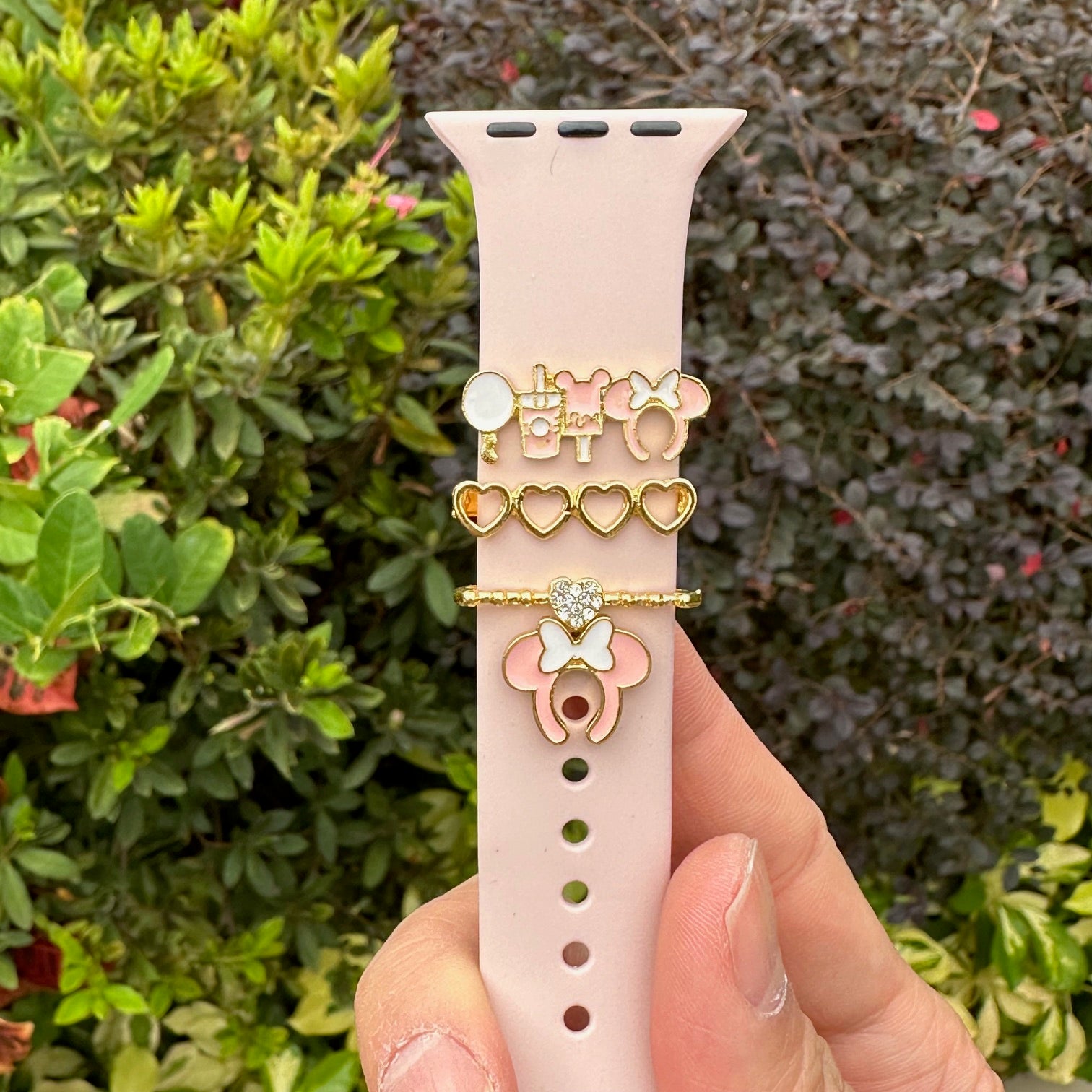 Pink Apple Watch band with decorative minnie mouse theme park snack charms held in front of a green bush
