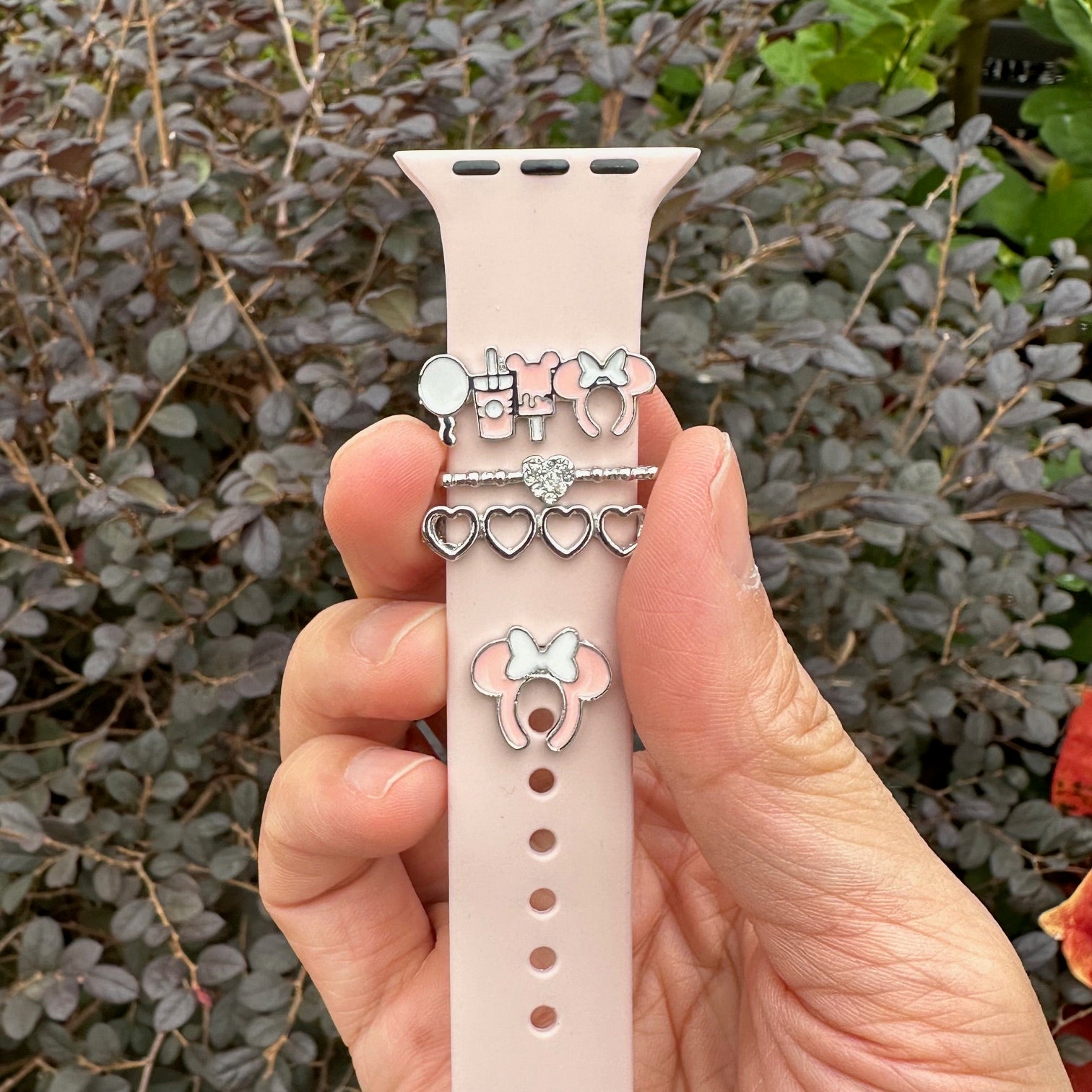 Hand holding a pink smartwatch band with decorative minnie mouse theme park snack charms against a leafy background