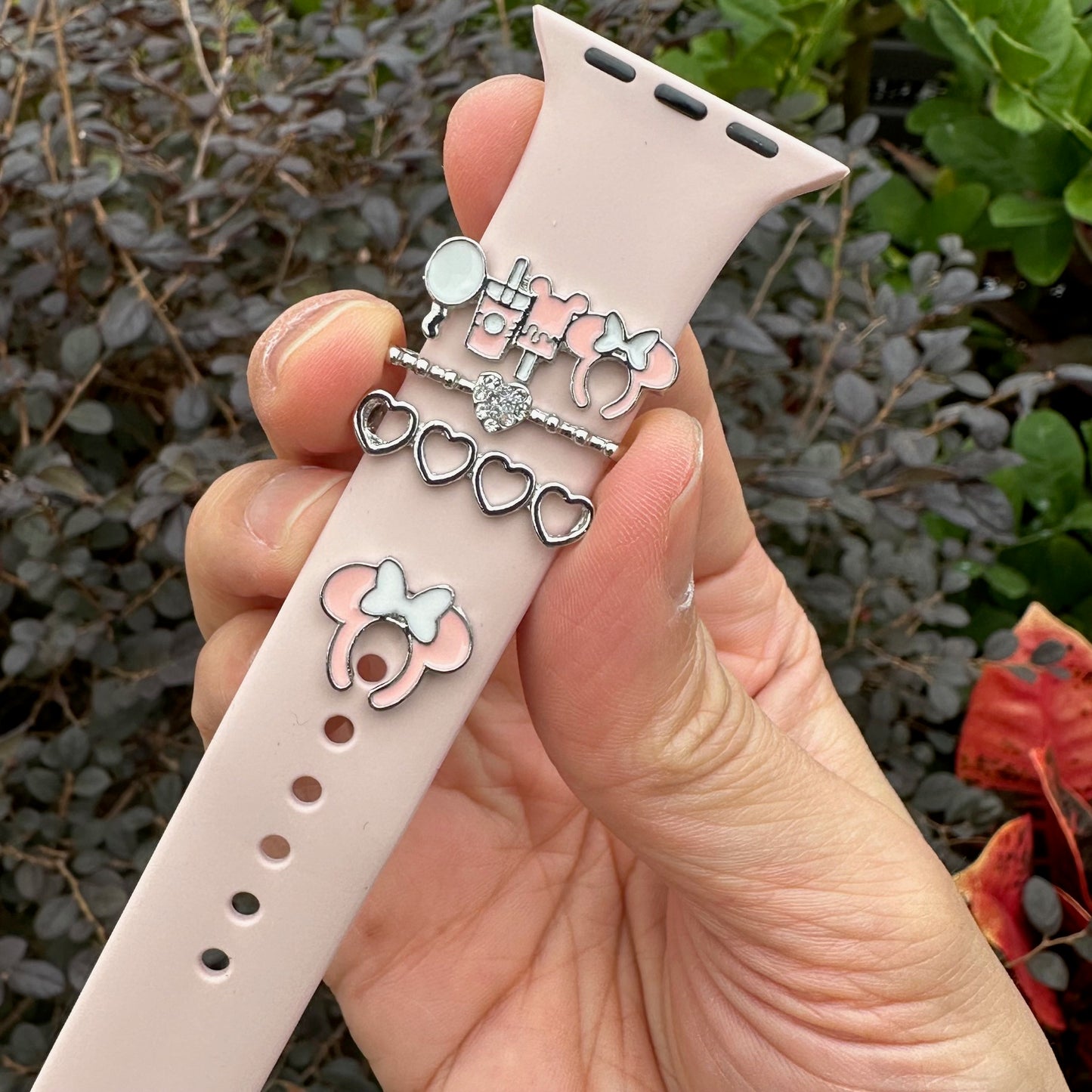 Hand holding a pink Apple Watch band with decorative minnie mouse theme park snack charms against a blurred natural background